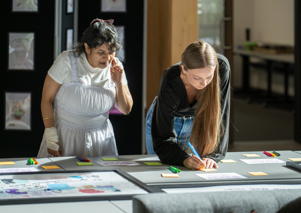 Two female presenting people stand in front of a table covered in yellow post-it notes. One, with long, loose blonde hair, in jeans and a loose black top, is writing. The other, with dark hair tied with a bow, in a pale dress and white t-shirt, is watching