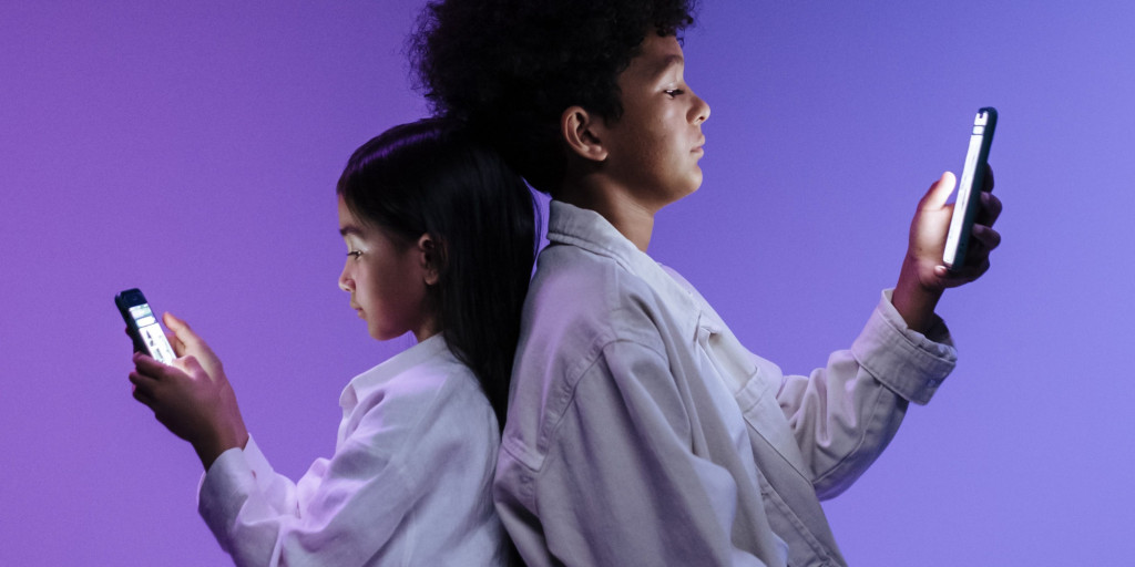 Two children, a girl and a boy, sitting back-to-back against a purple background, each absorbed in their own mobile phone.