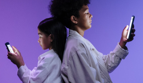 Two children, a girl and a boy, sitting back-to-back against a purple background, each absorbed in their own mobile phone.