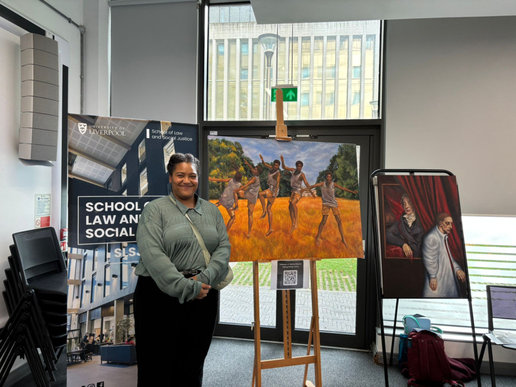 A woman stands in front of a poster and two artworks, one of people jumping in a field, another of a figure peering at another person from behind a curtain.