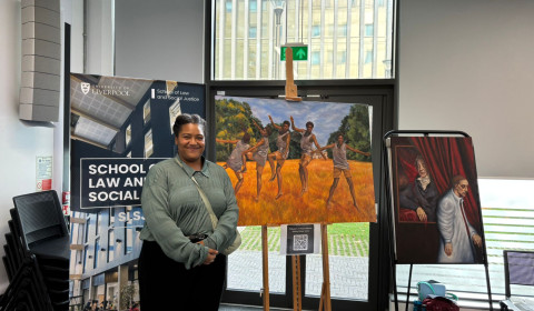 A woman stands in front of a poster and two artworks, one of people jumping in a field, another of a figure peering at another person from behind a curtain.