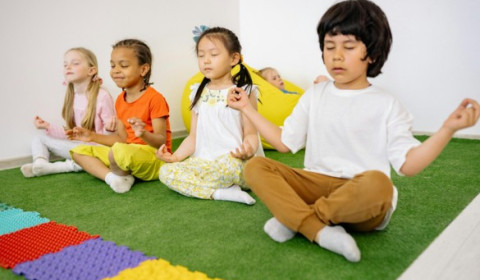 Four children sit in a row on a grass-like mat. They sit with their legs cross, eyes closed, and fingers pressed to thumbs.