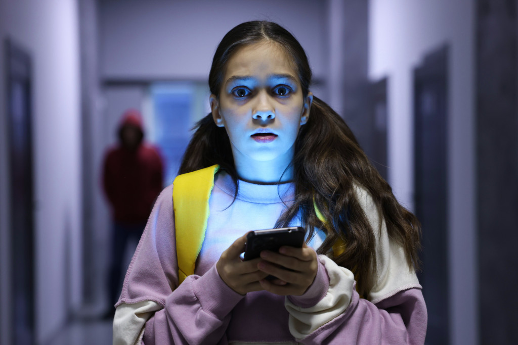 The picture shows a young girl standing in a dimly lit hallway while looking at her smartphone and a hooded preditor is in the background.