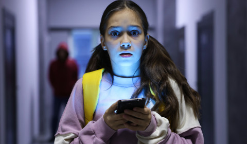The picture shows a young girl standing in a dimly lit hallway while looking at her smartphone and a hooded preditor is in the background.
