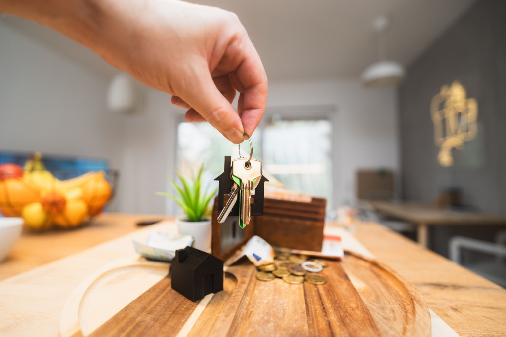 Hand holding a set of house keys attached to a house-shaped keyring above a wooden tray. On the tray are miniature house models, a wallet, coins, a bank card and a small plant. The background shows a bright, modern home interior with soft-focus furnishings