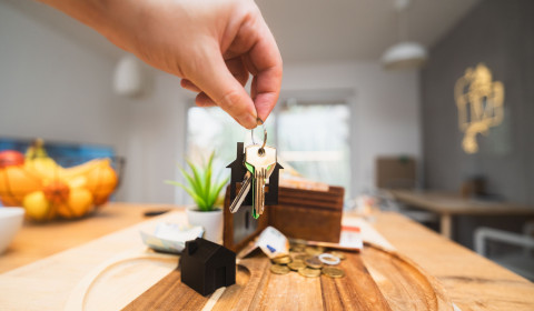 Hand holding a set of house keys attached to a house-shaped keyring above a wooden tray. On the tray are miniature house models, a wallet, coins, a bank card and a small plant. The background shows a bright, modern home interior with soft-focus furnishings