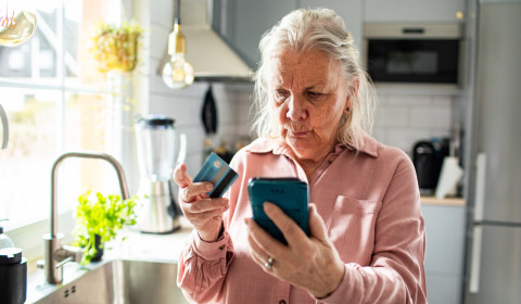 The picture shows an older woman standing in a bright, modern kitchen. She is holding a smartphone in one hand and a payment card in the other, looking closely at the phone with a serious, concentrated expression.